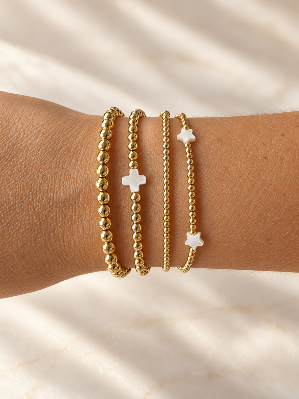Styling photo showing gold bead bracelets with white cross and star accents; bracelets sold separately