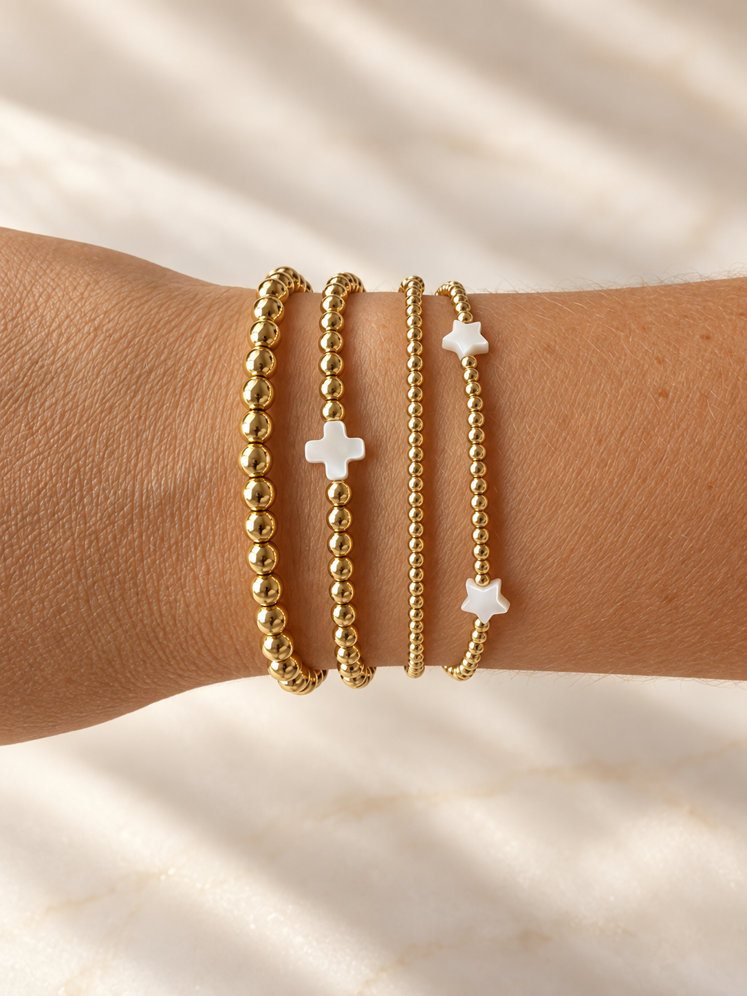 Styling photo showing gold bead bracelets with white cross and star accents; bracelets sold separately