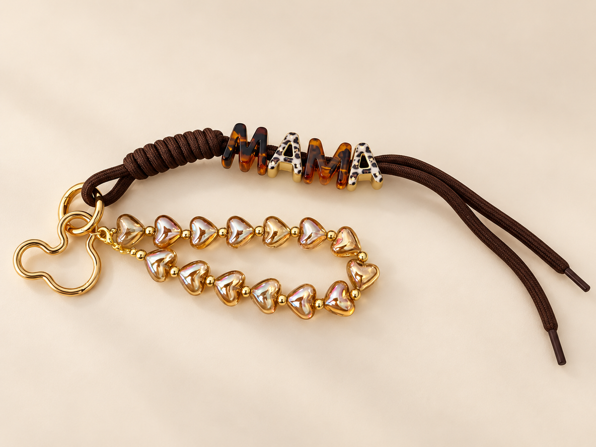 Brown cord MAMA bag charm with iridescent heart beads and gold-tone clasp on ivory background